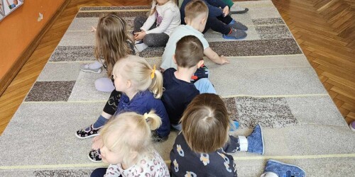 Dzieci siedzące w dwóch rzędach, dotykają się plecami